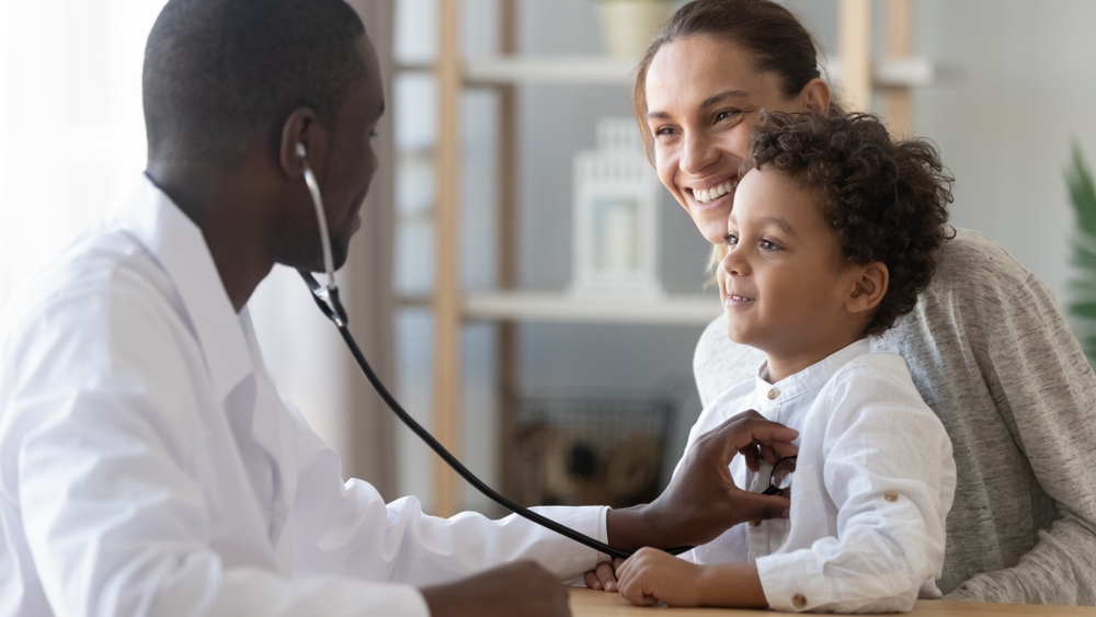 African male pediatrician holding stethoscope during preventive care visit with boy and mother